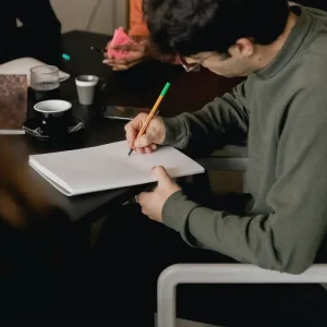 A person wearing glasses and a green sweater is seated at a table, writing in a notebook with a pencil. The scene includes other individuals in the background, partially visible, which enhances the sense of an engaging group setting. On the table next to them are a coffee cup and a glass. All appear to be diligently working on their Fair Housing Pre-Recorded material.