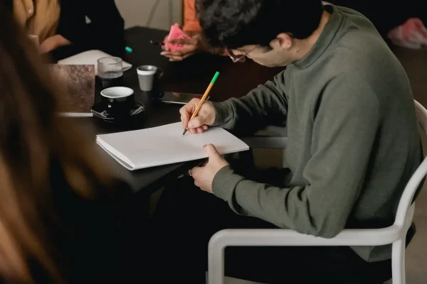 A person wearing glasses and a green sweater is seated at a table, writing in a notebook with a pencil. The scene includes other individuals in the background, partially visible, which enhances the sense of an engaging group setting. On the table next to them are a coffee cup and a glass. All appear to be diligently working on their Fair Housing Pre-Recorded material.