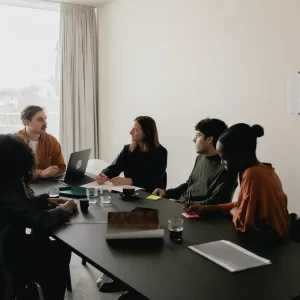 A group of six people are gathered around a black conference table in a bright, minimalist room. One person is using a laptop with the "Fair Housing Pre-Recorded" program open, and notebooks and glasses are scattered on the table. They appear engaged in a discussion, with notes pinned to the wall in the background.