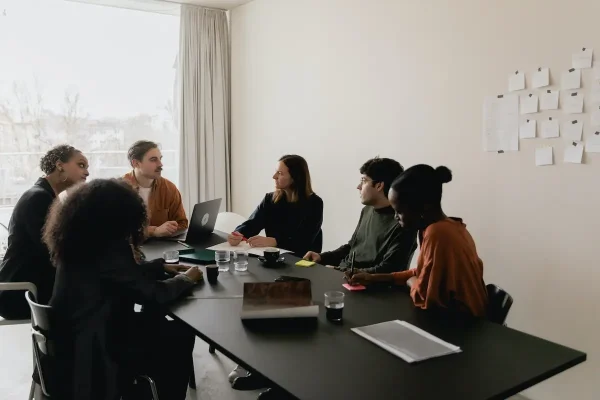 A group of six people are gathered around a black conference table in a bright, minimalist room. One person is using a laptop with the "Fair Housing Pre-Recorded" program open, and notebooks and glasses are scattered on the table. They appear engaged in a discussion, with notes pinned to the wall in the background.