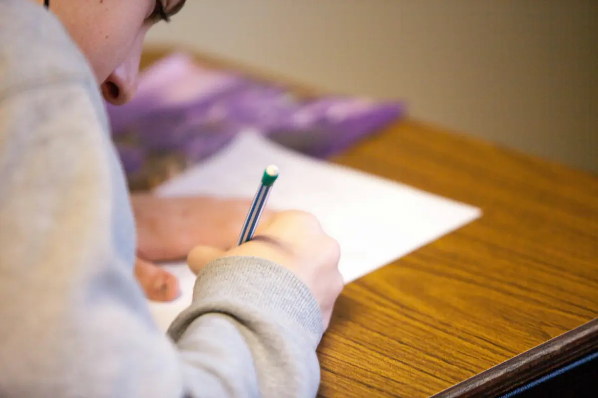 Close-up of a person wearing the C3P Pre-Recorded gray sweatshirt, holding a pencil, and writing on a sheet of paper at a wooden desk. The focus is on the hand and pencil, with the person's face partially visible. The background is blurred with some purple objects.