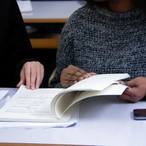 Two individuals are seated at a desk studying the book "Keeping in Compliance" together. One person is wearing a dark sweater with the letter "A" visible, while the other is in a light grey sweater. A smartphone and additional papers are placed on the desk between them.