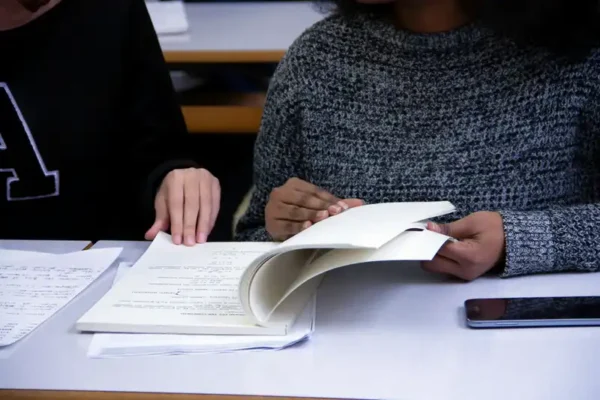 Two individuals are seated at a desk studying the book "Keeping in Compliance" together. One person is wearing a dark sweater with the letter "A" visible, while the other is in a light grey sweater. A smartphone and additional papers are placed on the desk between them.