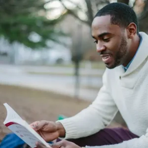 A man wearing a white sweater is seated outdoors on the ground, reading "Determining Income of School Employees" and smiling. The background shows trees and a pathway, creating a serene and peaceful atmosphere.