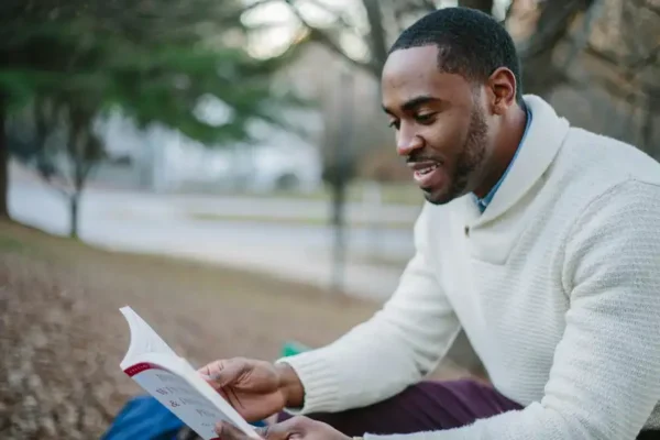 A man wearing a white sweater is seated outdoors on the ground, reading "Determining Income of School Employees" and smiling. The background shows trees and a pathway, creating a serene and peaceful atmosphere.