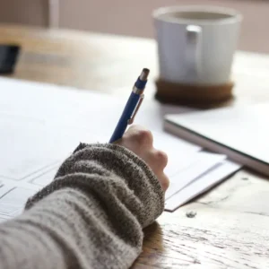 A person wearing a gray long-sleeve shirt writes with a blue-ink pen on sheets of paper spread on a wooden table. A white coffee mug and a closed copy of "Determining Income of School Employees" are situated in the background.