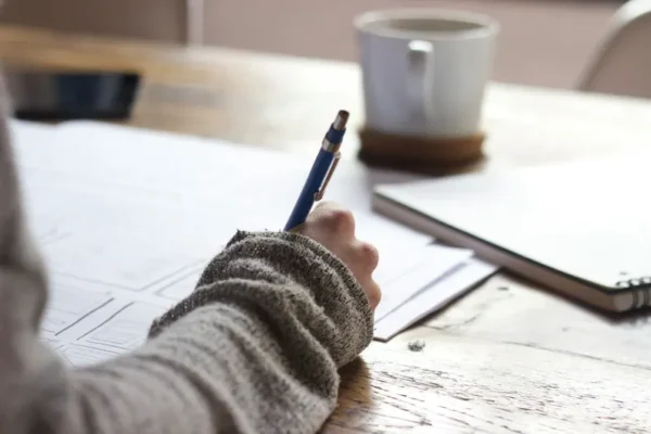 A person wearing a gray long-sleeve shirt writes with a blue-ink pen on sheets of paper spread on a wooden table. A white coffee mug and a closed copy of "Determining Income of School Employees" are situated in the background.